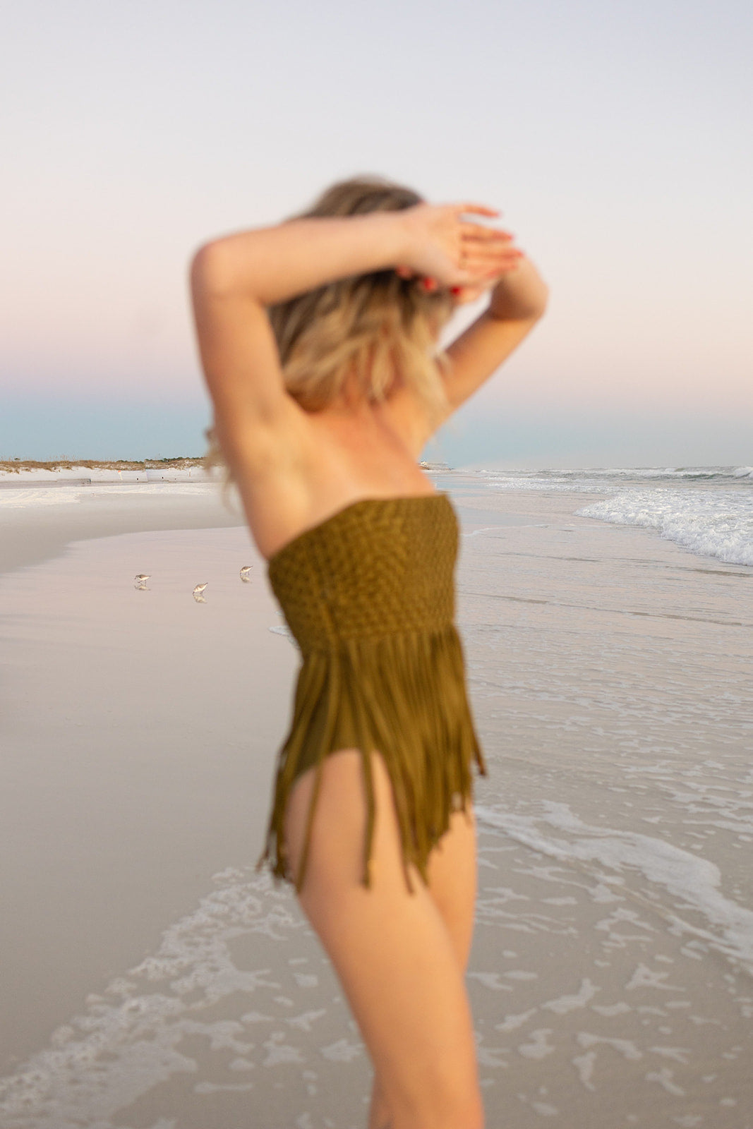 Blurred view of woman standing on the beach in an olive green strapless woven swimsuit with fringe details.