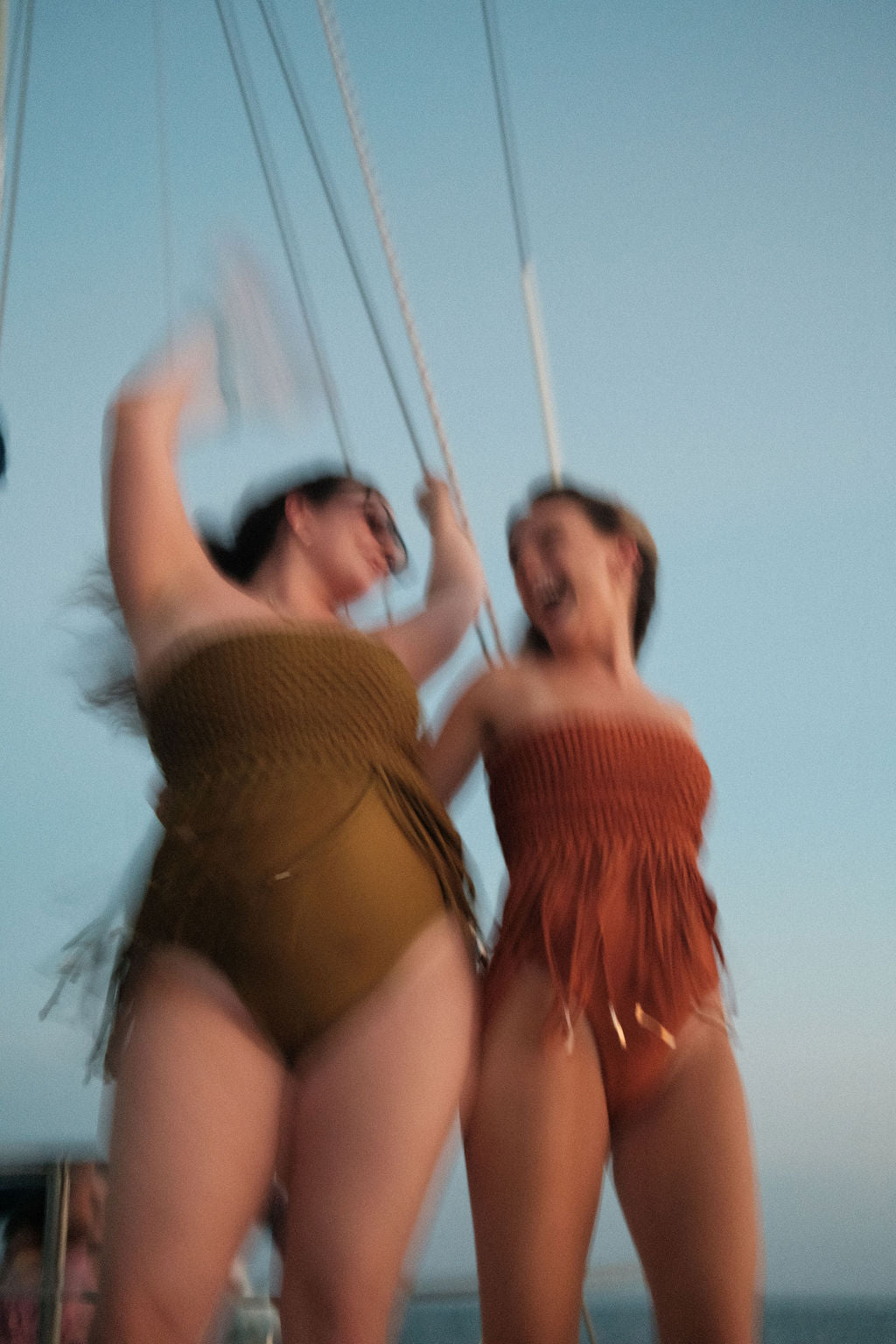 Two women dancing on a boat in woven fringe swimsuits—one in olive green, the other in rust orange.