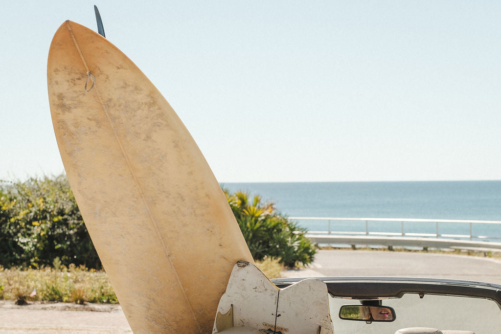 Surfboard resting in the back of a white convertible parked near the ocean, with clear skies and coastal greenery in the background.