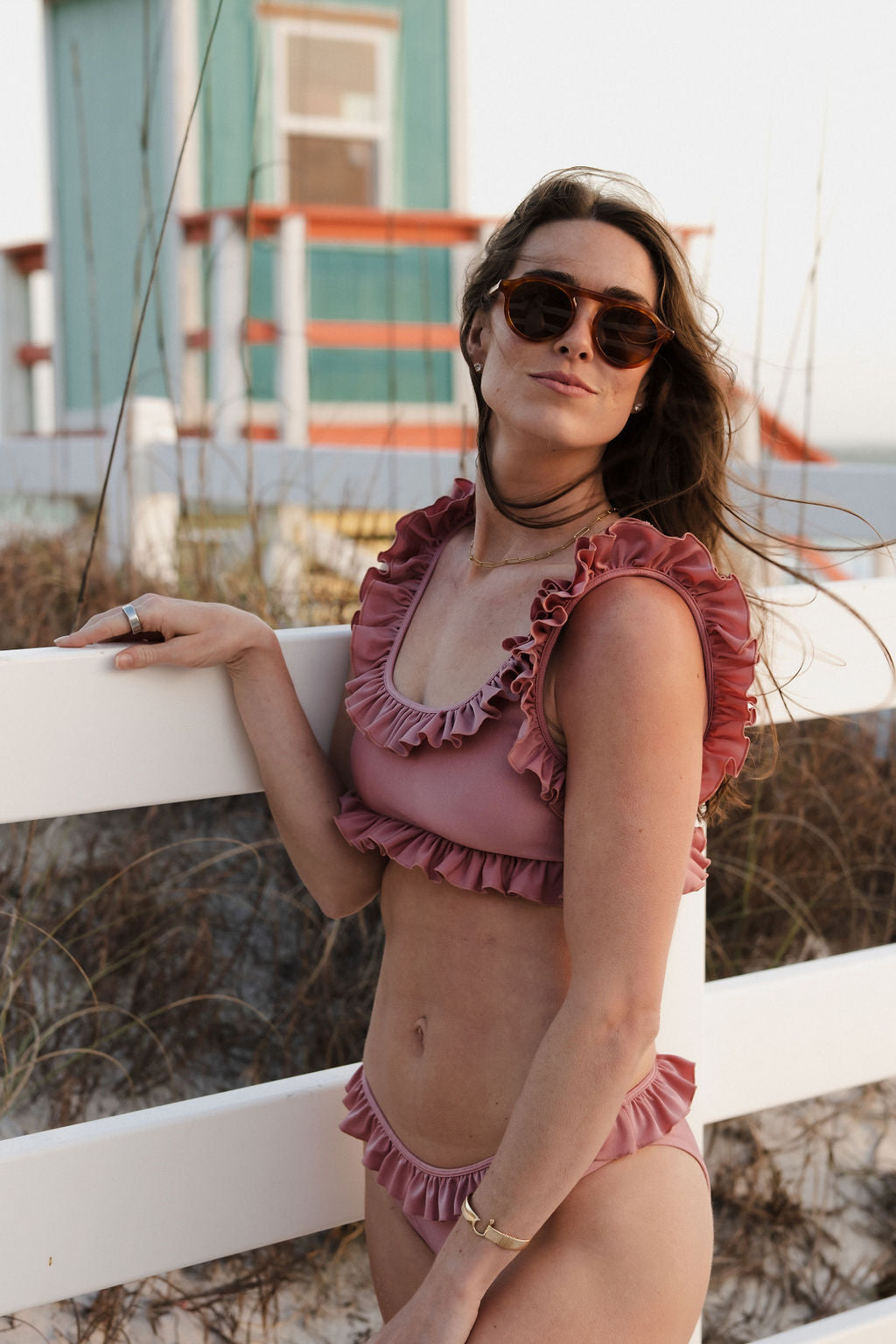 Confident woman in afterglow frill bikini with ruffle trim, leaning casually on white fence near beach house.